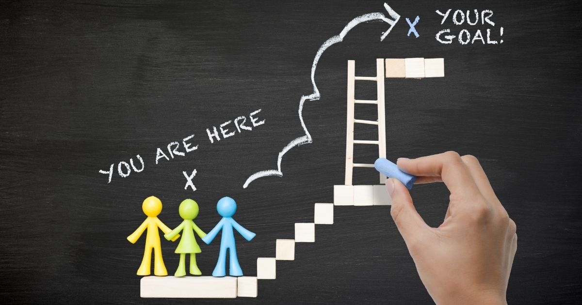 A hand draws a ladder on a blackboard with chalk, leading from stick figures labeled "You Are Here" to "Your Goal," symbolising progress and ambition.